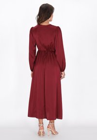 Bordowa sukienka z długim rękawem, dopasowaną talią, lejącym się dołem i miękką satynową teksturą. Charakteryzuje się bufkami na ramionach oraz okrągłym dekoltem.