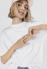 T-shirt bianco in cotone a maniche corte, abbinato a anelli d'oro sulle dita. Il modello ha i capelli biondi corti e indossa un berretto rosa chiaro.