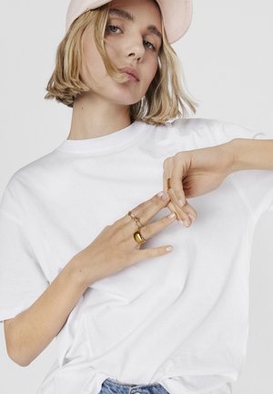 Camiseta de algodón blanca con mangas cortas, combinada con anillos dorados en los dedos. La modelo tiene el cabello corto y rubio, y lleva una gorra rosa claro.