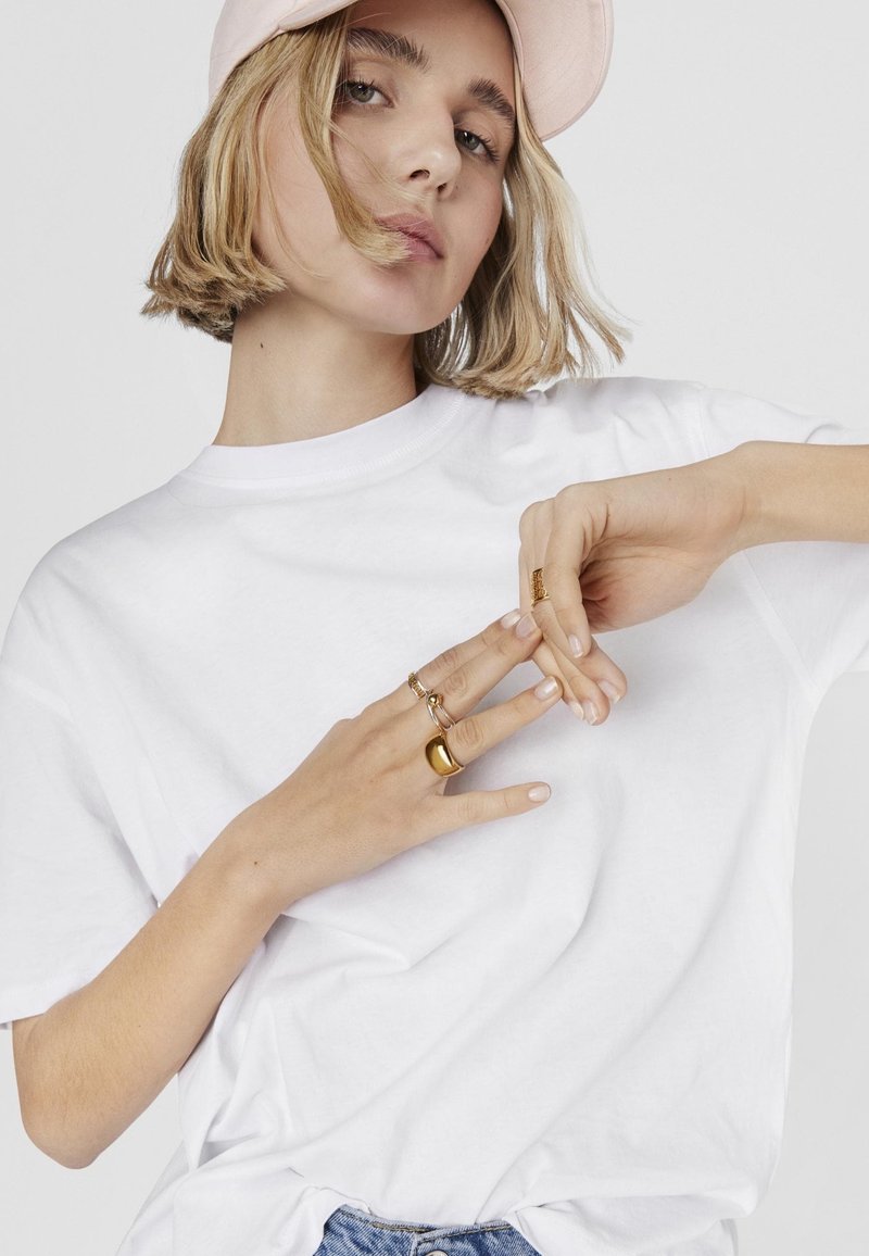 T-shirt bianco in cotone a maniche corte, abbinato a anelli d'oro sulle dita. Il modello ha i capelli biondi corti e indossa un berretto rosa chiaro.