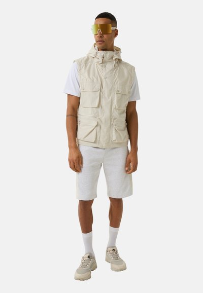 Beige ärmellose Weste mit mehreren Taschen, getragen über einem weißen T-Shirt, kombiniert mit grauen Shorts und beigen Turnschuhen. Reflektierende Sonnenbrille inklusive.