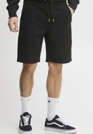 Black sweat shorts with an elastic waistband and drawstring, featuring side pockets. Worn with black and white sneakers and white socks.