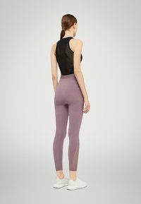 Zwarte mouwloze top met een textuurdesign, gecombineerd met nauwsluitende lila leggings met mesh zijpanelen. Model draagt witte sportschoenen.