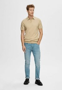Beige polo shirt met korte mouwen, gladde textuur, gecombineerd met lichtblauwe jeans, slim fit, omgeslagen aan de enkels, gedragen met zwarte schoenen.