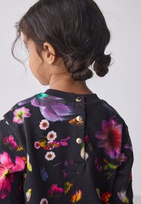 Mustast kangast lillemustriga pusa, millel on erksavärvilised lilled, kuldsed nupud selja taga ja ümar kaelus.