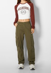 Wit en bordeauxkleurig cropped top met lange mouwen en "Melbourne" opdruk; olijfgroene cargobroek met meerdere zakken; gecombineerd met witte sneakers.