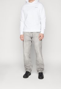 Felpa bianca con cappuccio e logo minimale, abbinata a jeans grigio chiaro e sneaker nere. Fit casual con una texture liscia.