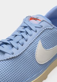 Ανοιχτό μπλε αθλητικό παπούτσι Nike με υφασμάτινη επάνω επιφάνεια με υφή, λευκό δερμάτινο λογότυπο, επίπεδα κορδόνια και μπεζ σόλα.