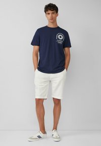 Dunkelblaunes Baumwoll-T-Shirt mit Druck auf der Vorderseite, kombiniert mit weißen Shorts und beigen Sneakers mit blauen Akzenten.