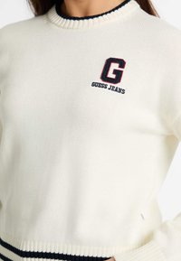 Krém színű pulóver bordás kerek nyakkal és navy csíkozott hem.Jellemzői a kék és piros színben hímzett "G" logó és a "GUESS JEANS" felirat.Texturált kötött anyag.