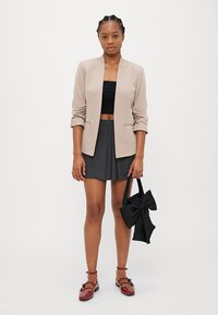 Beige sakový blejzer s vyhrnutými rukávmi, čierny crop top a šedá sukňa s vysokým pásom. Doplnky tvoria čierna mašľa na kabelke a červené balerínky.
