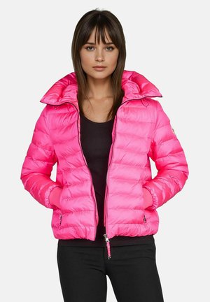 Frau mit glattem braunem Haar trägt eine leuchtend pinke Steppjacke über einem schwarzen Oberteil, Hände in den Taschen der Jacke, vor einem schlichten Hintergrund.