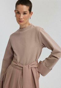 Dlhý rukáv, top v odtieni mauve s vysokým golierom a širokými manžetami, s viazaným pásom v rovnakom odtieni. Hladká tkanina a prispôsobený strih.