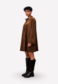 Frau mit kurzem, lockigem Haar, die ein lockeres braunes Kleid und hohe schwarze Lederstiefel trägt, steht in neutraler Pose vor einem weißen Hintergrund.