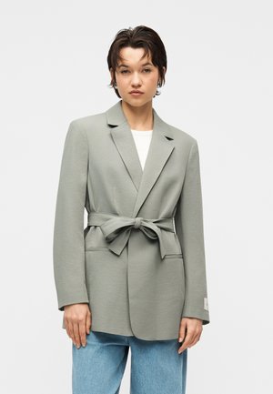 Mladý človek s krátkymi tmavými vlasmi, oblečený v svetlosivom saku so zaväzovaním v páse cez bielu košeľu a modré rifle, stojaci pred jednoduchým pozadím.