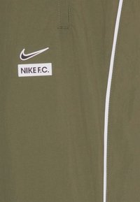 Tissu vert olive avec le logo Nike swoosh blanc au-dessus du texte "NIKE F.C." et une bande verticale blanche sur le côté droit.
