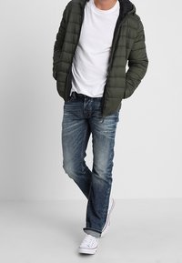 Veste matelassée vert olive avec fermeture éclair, associée à un jean en denim bleu et des baskets blanches. La veste est dotée d'une capuche et d'une texture matelassée.