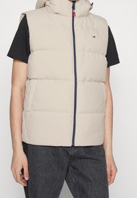 Beige pufferväst med huva, dragkedjestängning och marinblå detaljer. Har en kviltad textur och sidofickor. Bärs över en svart t-shirt.