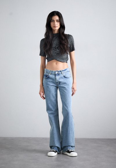 Grå kort topp med kort ärm, matchad med ljusblå utsvängda jeans. Modellen bär svarta och vita sneakers. Enkelt bakgrund.