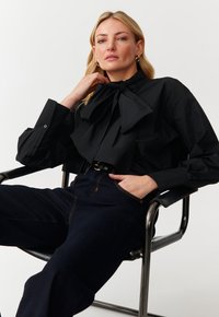 Blusa nera con un grande fiocco al collo, maniche lunghe con pliche, abbinata a pantaloni in denim scuro. Modella seduta su una sedia.
