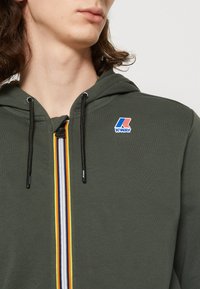 Felpa con zip color verde oliva, dotata di una zip frontale e strisce arancioni, gialle e blu; cordini neri; logo K-Way sul petto.