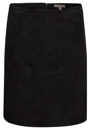 Femme portant un manteau texturé couleur rouille, un pull à col roulé marron, une jupe noire et des bottes noires montant jusqu'au genou sur un fond uni.