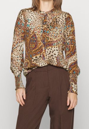Woman wearing long-sleeve blouse with colorful paisley and leopard print, paired with high-waisted brown trousers, standing against a plain background.