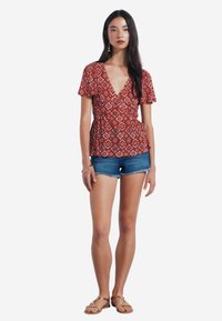 Rote gemusterte Bluse mit kurzen Ärmeln, V-Ausschnitt und dekorativen Knöpfen, kombiniert mit Jeansshorts und Sandalen.