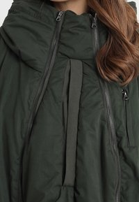 Tmavě zelená zateplená bunda s vysokým límcem, několika svislými zipy a dlouhými vlnitými hnědými vlasy viditelnými na pravé straně.