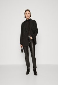 Blazer dublu din material cu dungi negre peste un top tricotat negru, asortat cu pantaloni din piele negri și cizme până la glezne; ținând o geantă mică.