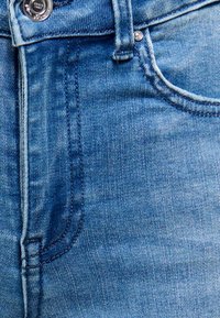 Gros plan sur un jean en denim bleu montrant le bouton, la zone de la fermeture éclair, la poche avant et les détails visibles de couture.