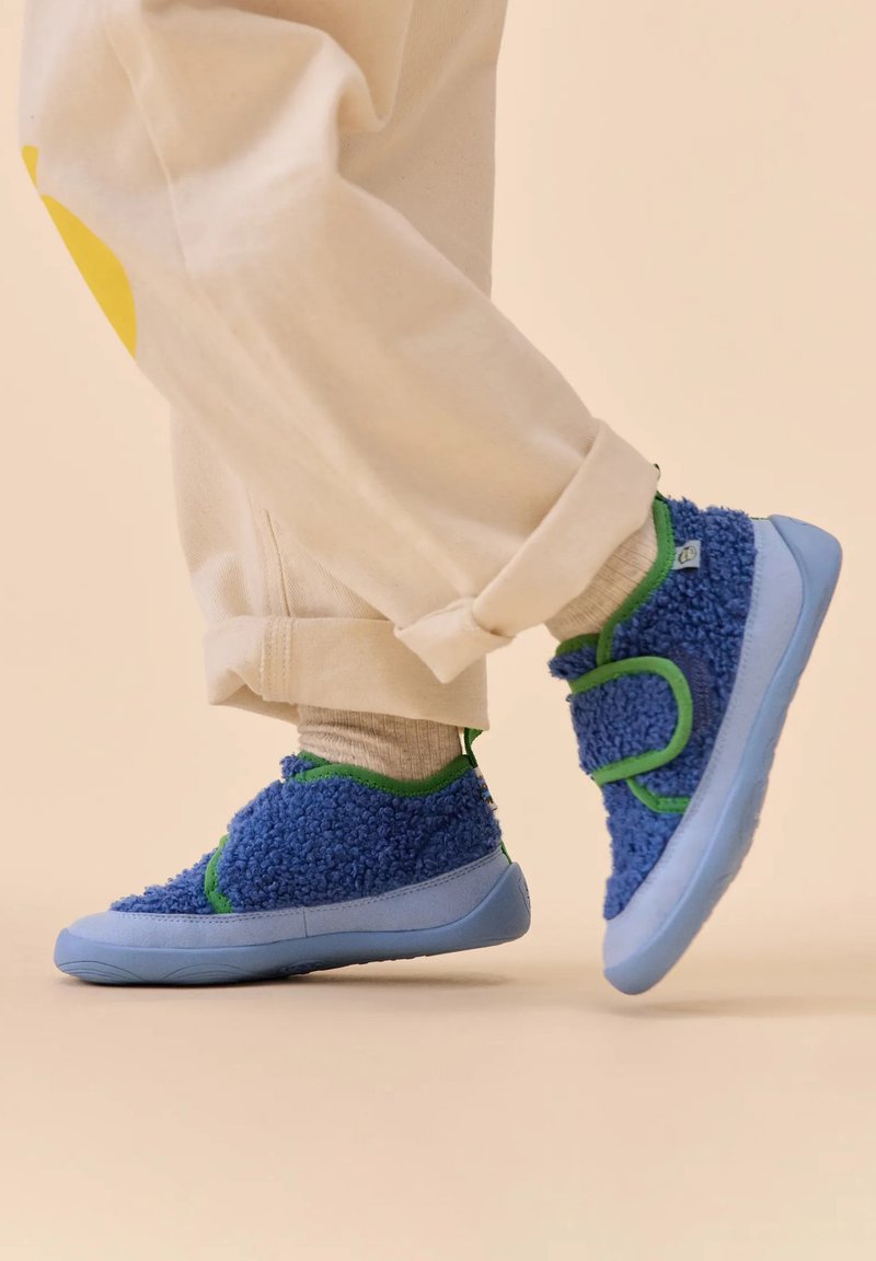 Blaue, fuzzy Schuhe aus strukturiertem Stoff mit grünen Akzenten; verfügen über einen Klettverschluss und eine hellblaue Sohle; getragen mit gerollten, hellen Hosen.