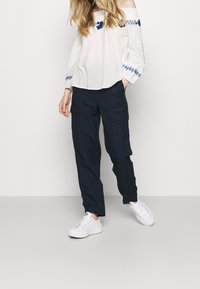 Biała bluzka odkrywająca ramiona z niebieskimi haftami kwiatowymi, zestawiona z granatowymi spodniami cargo i białymi trampkami. Swobodny strój z fakturowanej tkaniny.