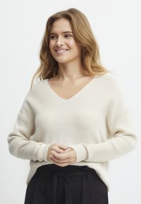 Pull en tricot beige avec un col en V, texture côtelée et coupe ample. Manches longues avec détails aux poignets, associé à un pantalon noir taille haute.