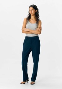 Femme aux longs cheveux bouclés, se tenant les bras croisés, portant un débardeur gris, un pantalon bleu marine et des sandales à talons noirs.
