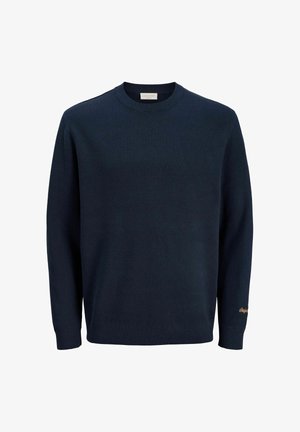 Uśmiechnięty chłopiec w beżowej czapce, granatowej bluzie, jasnoniebieskich szerokich dżinsach i beżowych butach sznurowanych stoi na jednolitym jasnym tle.