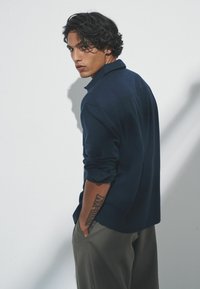 Granatowa koszula z kołnierzykiem, długie rękawy podwinięte do łokci, wykonana z lekkiego materiału, prezentowana na modelu z tatuażem na ramieniu.