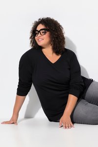 Femme aux cheveux bouclés avec des lunettes noires portant un haut noir à manches longues et un pantalon gris, s'appuyant sur une surface blanche sur un fond blanc.