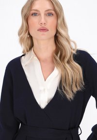 Marineblaues Wickelkleid mit langen Ärmeln, aus weichem Material gefertigt, mit tiefem V-Ausschnitt und gebundenem Taillenbereich. Darunter eine cremefarbene Satinbluse mit Kragen.