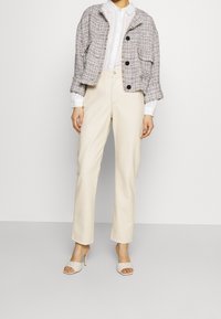 Texturovaný béžový tweedový kabát s velkými černými knoflíky, bílá košile s límečkem, krémové kalhoty a světlé sandály na podpatku s pásky.