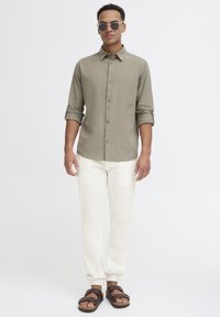 Olive green button-up shirt, rolled sleeves, paired with white elastic cuffed pants and brown sandals, set against a plain background.