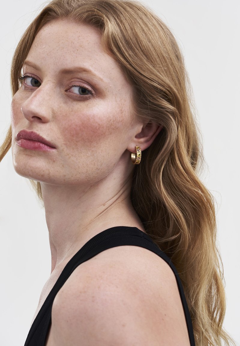 Aukso hoop auskarai su sudėtingu raštu, lygiomis tekstūromis ir apvalia forma, dėvimi modelio su banguotais, šviesiai rudais plaukais.