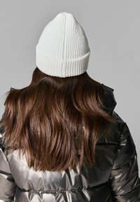 Vit stickad mössa med ribbad textur, rundad krona och vikbar skärm, bärs på långt, vågigt brunt hår, mot en grå bakgrund.