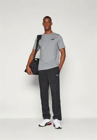 Tânăr în picioare, îmbrăcat cu un tricou gri Puma, pantaloni de trening negri Puma, adidași albi și purtând o geantă de sport neagră pe umăr.