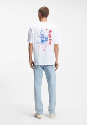 Man draagt een wit T-shirt met blauwe cocktailglazen en rode tekst "Happy Hour Every Hour" op de achterkant, gecombineerd met lichtblauwe jeans en witte schoenen.