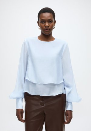 Gaiši zilā blūze ar slāņotu dizainu, garām pūkainām piedurknēm un apaļu kakla izgriezumu. Valkāta kopā ar brūnām ādas tipa biksēm. Mīksts, plūstošs audums.