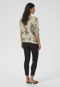 Blusa a stampa floreale in beige chiaro con accenti verdi e neri, con maniche a tre quarti e una vestibilità rilassata, abbinata a pantaloni neri slim-fit.