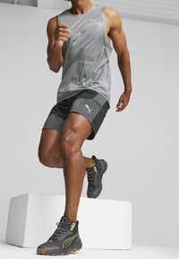 Top senza maniche grigio con motivo e pantaloni corti neri con logo bianco; indossa scarpe da ginnastica grigie e nere con accenti gialli; posa a metà passo.