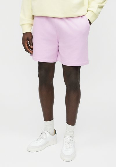 Mandlig model iført lyse lilla shorts, hvide sokker, hvide sneakers og en lysegul sweatshirt, stående med den ene hånd i lommen.