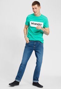 T-shirt di cotone turchese con logo "Wrangler" bianco, abbinata a jeans in denim blu e sneakers nere. Outfit casual con una vestibilità rilassata.